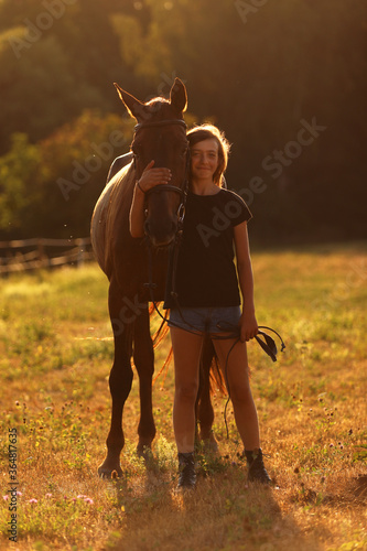 Young girl with chestnut horse on meadow during summer sunset. Romanic scene