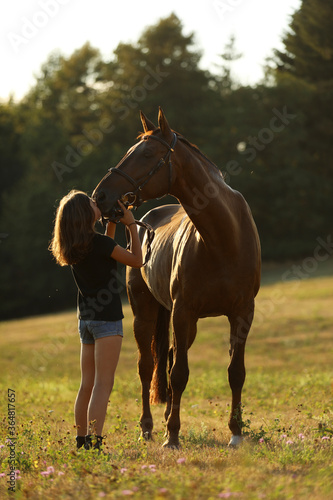 Teenaged girl kissing chestnut horse on pasture. Summer sunset on meadow, romantic scene