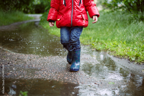 a boy in a red jacket and blue boots is walking along an asphalt path among puddles, dripping rain, in the background is green grass. lmage with selective focus