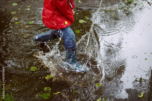a boy in blue jeans, patterned boots and a red jacket during the rain jumps in a large puddle, splashes fly.
lmage with selective focus photo