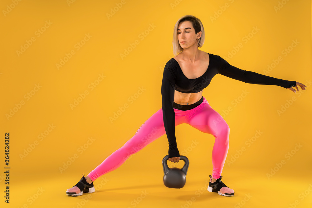 Fitness woman in fashionable pink and black sportswear work out with kettlebell on yellow background. Strength and motivation.