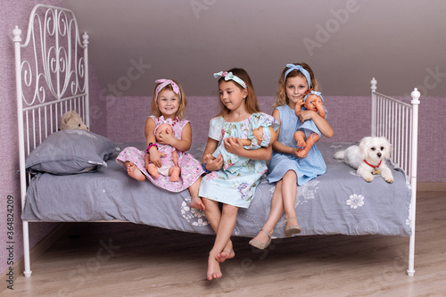 three little girls-sisters with loose hair and dresses sitting on the bed in the bedroom holding dolls in their hands