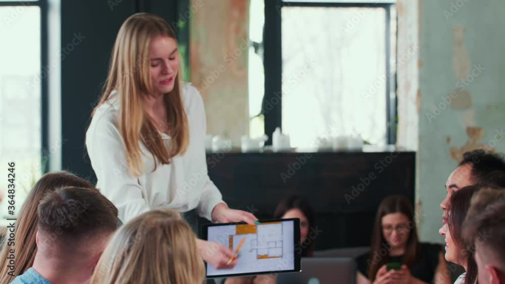 Attractive young gen-z happy Caucasian blonde CEO business woman gives presentation with digital tablet at team meeting. Attractive young gen-z happy Caucasian blonde CEO business woman gives presentation with digital tablet at team meeting.