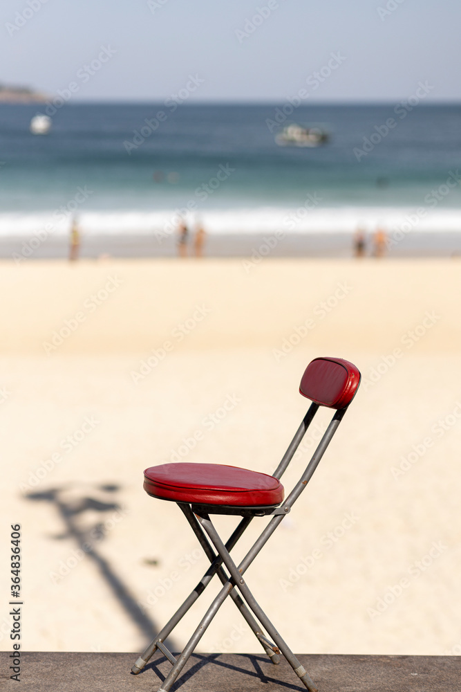 Small red chair in sunlight with out of focus blurred the beach of Ipanema, shadow of lamppost and sunbathers in the background Small red chair in sunlight with out of focus blurred the beach of Ipanema, shadow of lamppost and sunbathers in the background