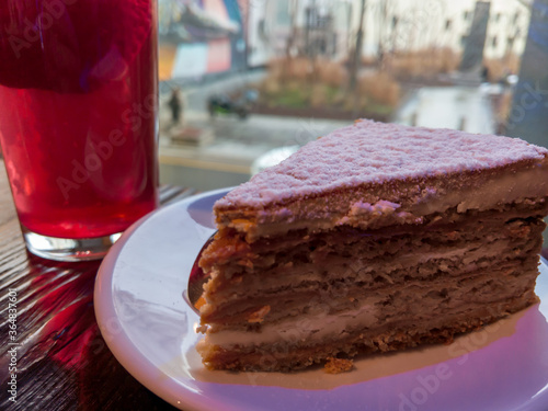 Close-up of a slice of honey cake on a white saucer standing on a rough wooden table with strawberry compote in the background Maya Plisetskaya Park photo