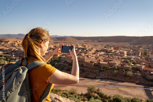 Woman photographing village with smart phone while standing against clear sky on sunny day