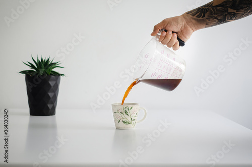 Mano de hombre vertiendo café recién hecho en una taza sobre una mesa blanca, en segundo plano una maseta negra con una planta. Proceso de preparación de café colombiano.  photo