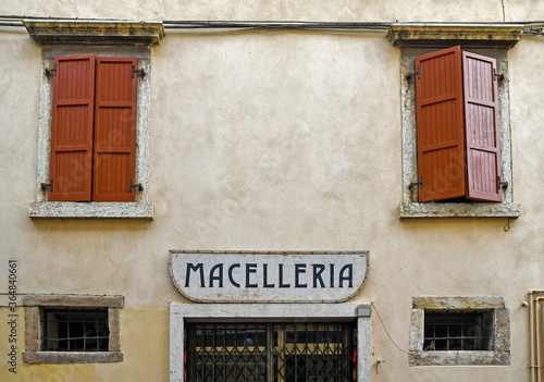 Exterior of an old building with closed wooden shutters, stone frames, and a rusty metal shop sign that says: 