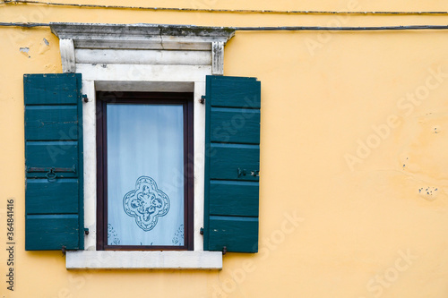 Close-up of the window of an old building with an embroidered tent, stone frame and green wooden shutters on a yellow wall, Italy