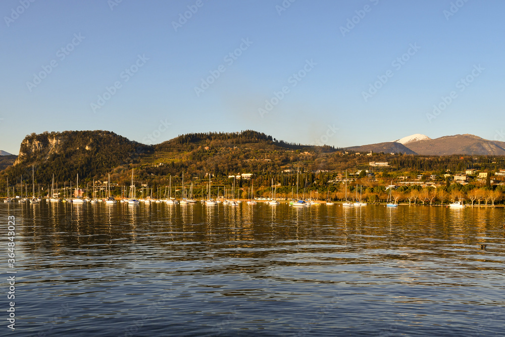 Scenic view of Lake Garda with moored boats and the mountainous coast in the background in a sunny winter day, Veneto, Italy
