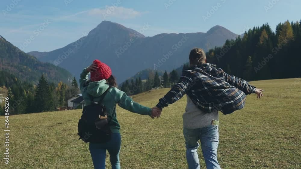 Back of two guys holding hands with backpacks on summer vacation or holidays running through alpine meadows. Silhouette of young family of travelers spending time together outdoors. Concept of enjoy. Back of two guys holding hands with backpacks on summer vacation or holidays running through alpine meadows. Silhouette of young family of travelers spending time together outdoors. Concept of enjoy.