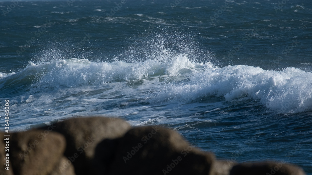 White wave on ocean surface, on a blustery, windy day with rocks in foreground.