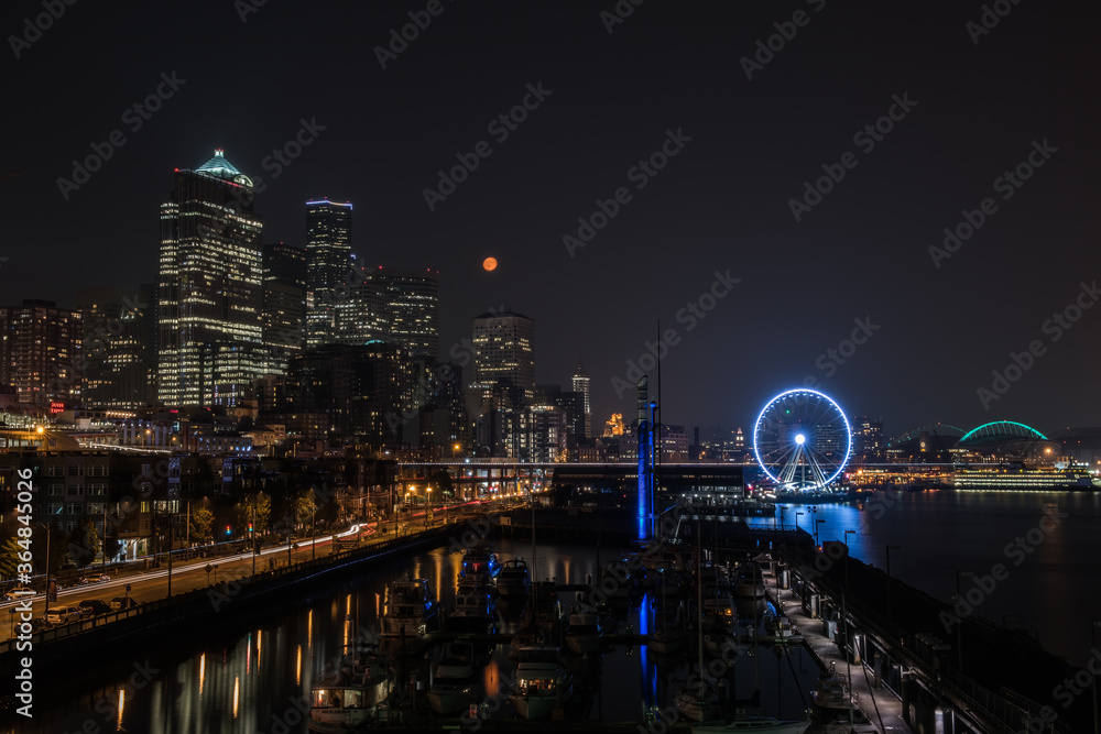 Downtown Seattle at night with a full moon, waterfront with reflections and a blue ferris wheel.