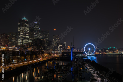 Downtown Seattle at night with a full moon, waterfront with reflections and a blue ferris wheel.