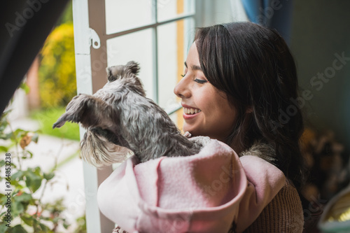 Girl looking out the window carrying her pet in her arms, dressed in a pink sweater, her dog Schnauzer enjoys the company of her friend, an afternoon of relaxation and company of her pet. 3