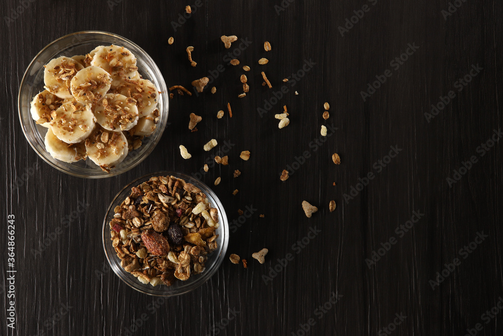 Bananas cut into slices with granola and cereal on dark gray wooden background in glass bowls on a table. Top view, copy space Bananas cut into slices with granola and cereal on dark gray wooden background in glass bowls on a table. Top view, copy space