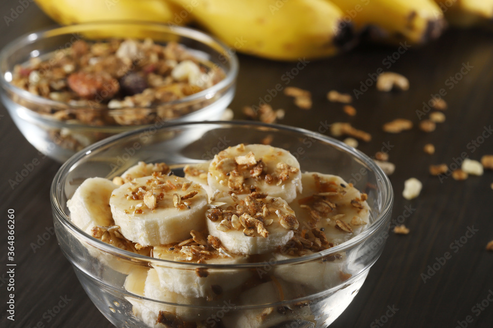 A banquet of bananas with granola and cereal on dark gray wooden background and a sliced banana in a glass bowl on a table. Selective focus A banquet of bananas with granola and cereal on dark gray wooden background and a sliced banana in a glass bowl on a table. Selective focus