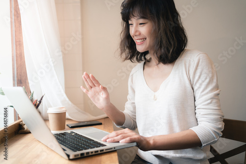 Young Asian woman working and video conferencing with laptop computer. A happy woman with smiley face working from home. A cup of coffee on table. Work from home concept