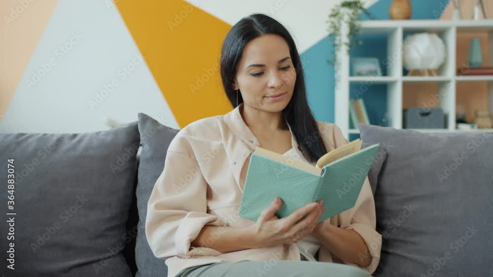 Good-looking young woman is reading book and smiling then turning page sitting on comfortable sofa alone in nice apartment. Youth, literature and lifestyle concept. Good-looking young woman is reading book and smiling then turning page sitting on comfortable sofa alone in nice apartment. Youth, literature and lifestyle concept.