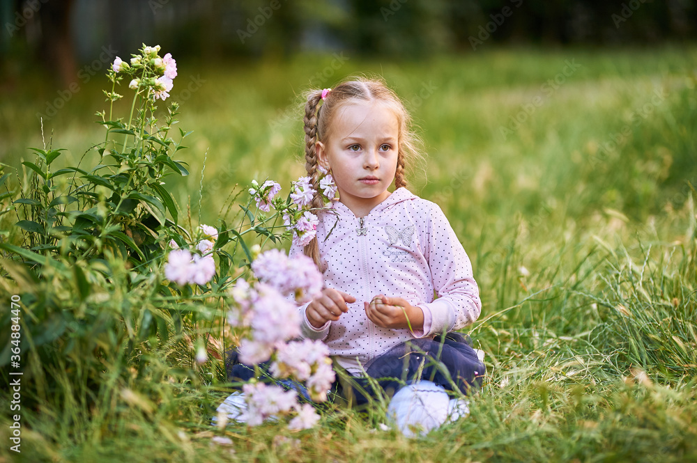 The little girl sits on the grass near the bush with pink flowers and looks to the side The little girl sits on the grass near the bush with pink flowers and looks to the side