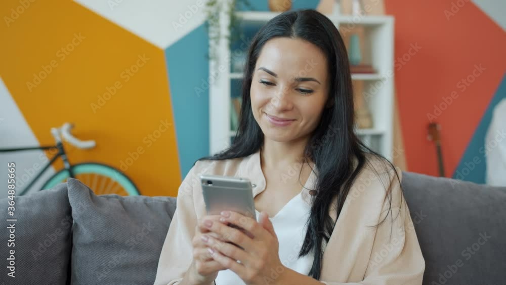 Attractive young woman is holding smartphone touching screen and smiling relaxing at home sitting on sofa in cozy room. Modern technology, internet and youth concept. Attractive young woman is holding smartphone touching screen and smiling relaxing at home sitting on sofa in cozy room. Modern technology, internet and youth concept.