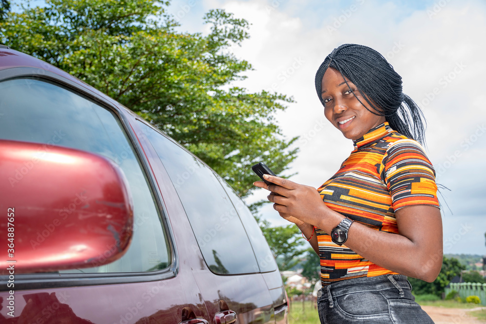 a young black woman standing close to a vehicle, smiling and using her mobile phone