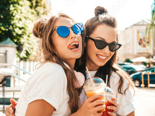 Two young beautiful smiling hipster girls in trendy summer clothes. Carefree women posing outdoors.Positive models holding and drinking fresh cocktail smoothie drink in plastic cup with straw