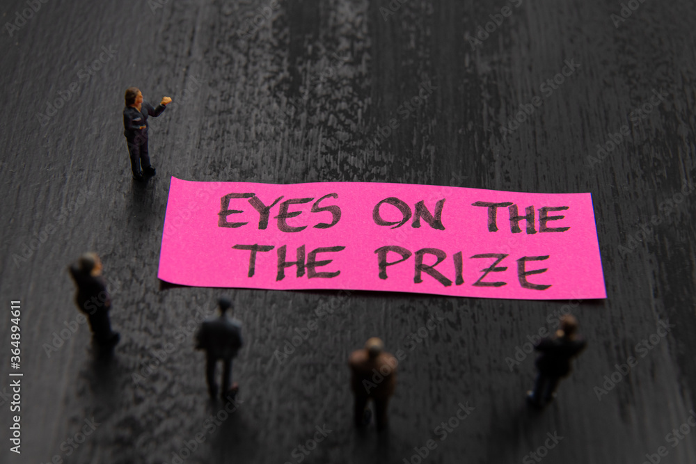Strategy meeting with miniature figurines posed as business people standing around post-it note with Eyes On The Prize message, minimalist abstract concept image with focus on text