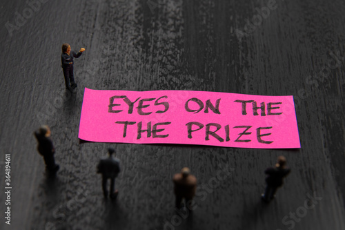 Obraz na płótnie Strategy meeting with miniature figurines posed as business people standing around post-it note with Eyes On The Prize message, minimalist abstract concept image with focus on text w salonie
