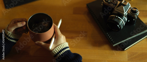 Female freelancer holding coffee mug while sitting at workspace with diary notebooks and camera