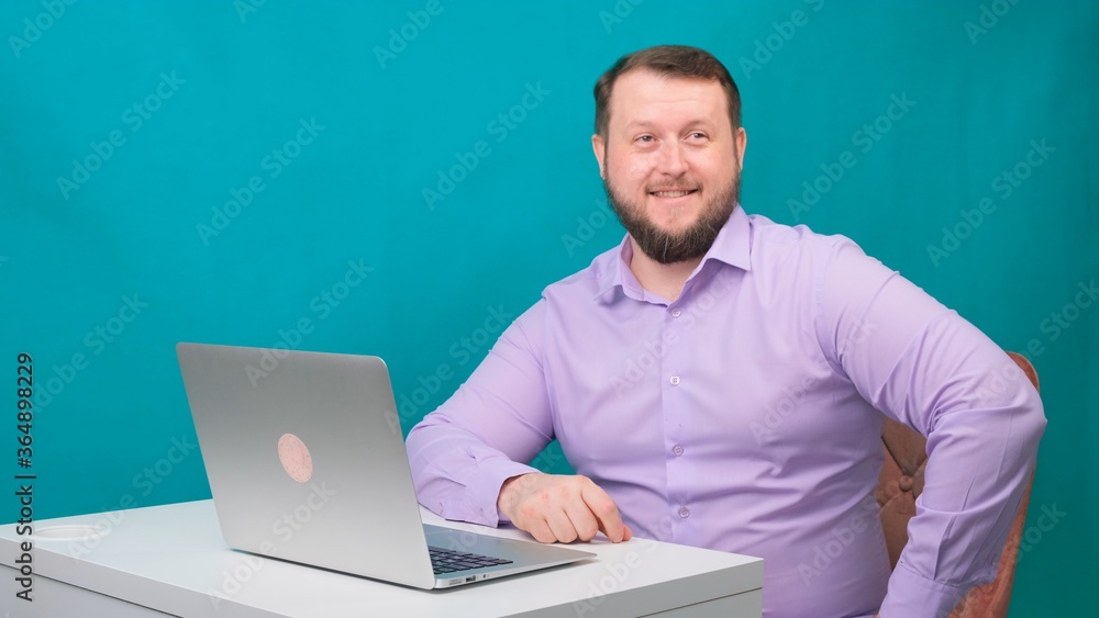 Young happy businessman smiling and speak into the camera. Portrait of a male laughing while looking at his laptop. Man working at his desk in the office