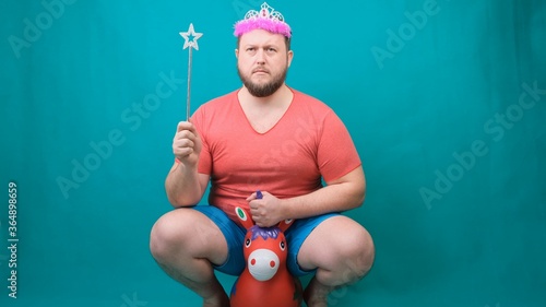 handsome bearded freaky man in a pink T-shirt with a deadema on his head is saddened riding a unicorn with a magic wand in his hand. A funny wizard joke to make and fulfill a wish. photo