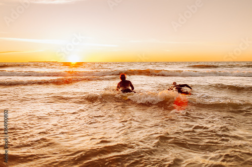 Two young male surfers in black wetsuits with longboards in a water at sunset ocean. Water sport adventure camp and extreme swim on summer vacation. © Olga