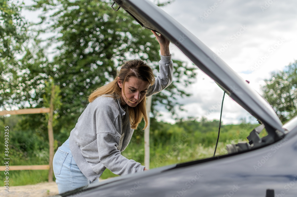 Young charming girl stands near the open hood of a broken car trying to repair it Young charming girl stands near the open hood of a broken car trying to repair it