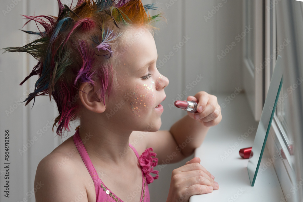 Obraz premium A little girl is getting ready for a party. A child paints her lips in front of a mirror. The girl is dressed in an evening pink dress.
