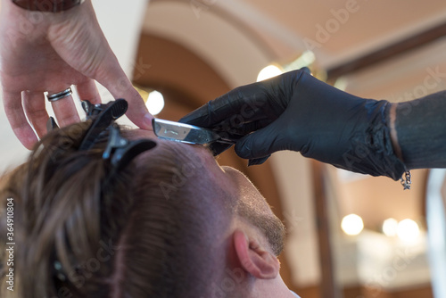 barber shaving a young bearded barber shop customer guy with a razor. close up. beard haircut, dangerous razor, professional hair and beard care.