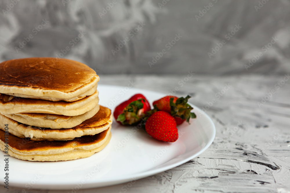 A stack of pancakes with a pink tea mug and fresh strawberries on a white plate on a light background. Horizontal orientation. Homemade american pancakes. Copy space. A stack of pancakes with a pink tea mug and fresh strawberries on a white plate on a light background. Horizontal orientation. Homemade american pancakes. Copy space.