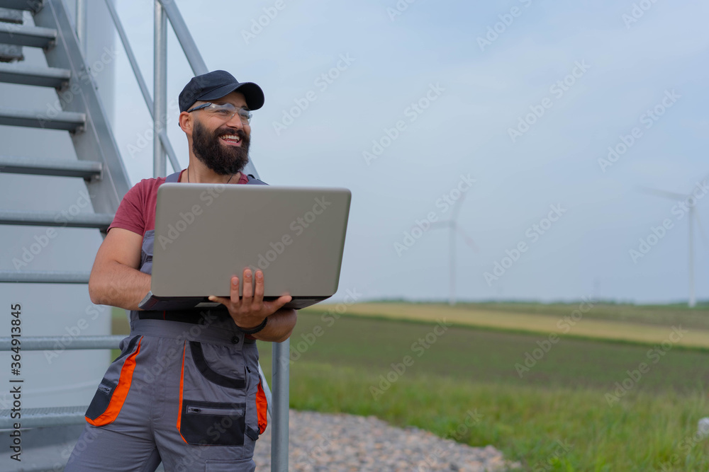 Young worker with strong beard in front of wind turbine. Working on laptop. 