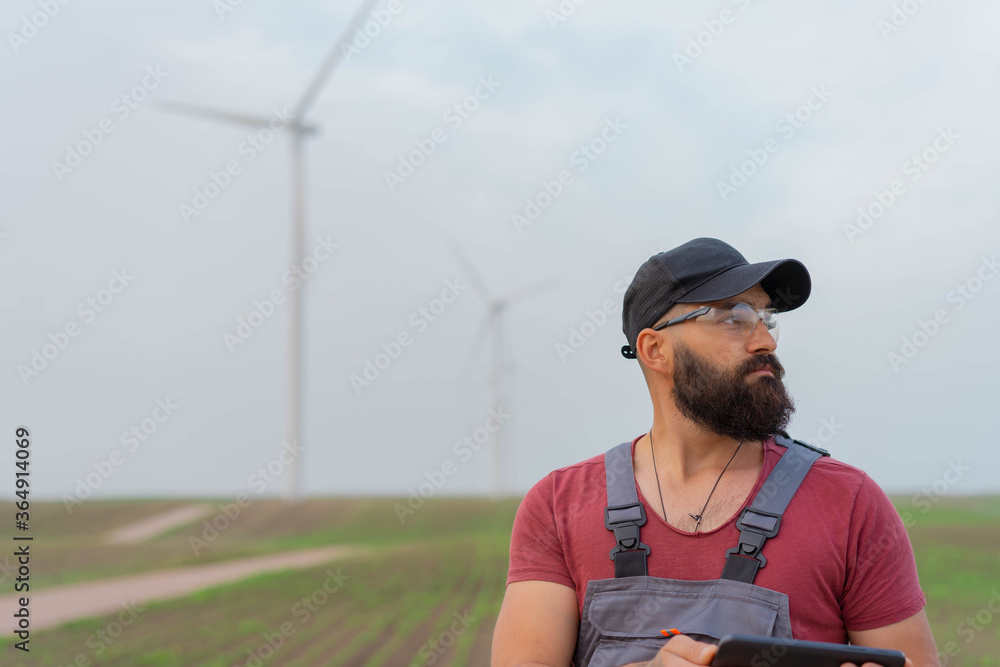 Young worker with strong beard in wind park. Holding tablet. Wind Mill in background. Young worker with strong beard in wind park. Holding tablet. Wind Mill in background.