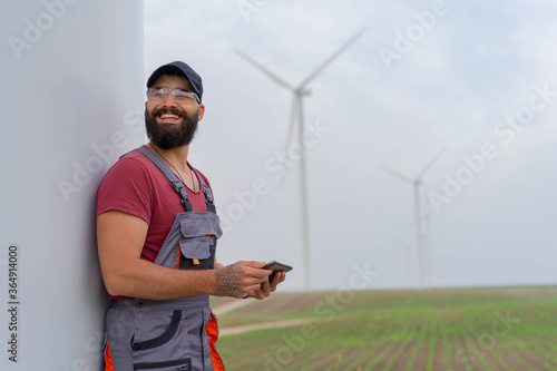 Young worker with strong beard in wind park. Holding smart phone. Wind Mill  in background. photo