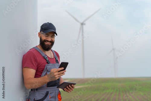 Young worker with strong beard in wind park. Holding smart phone and tablet. Wind Mill  in background.