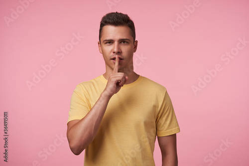 Indoor photo of young brunette guy dressed in casual wear keeping raised index finger on his mout while looking at camera, isolated over pink background