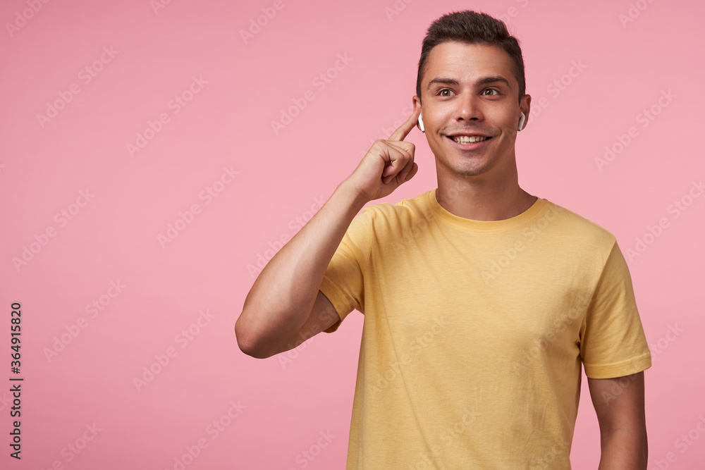Happy young brown-eyed brunette male smiling cheerfully while listening to music and keeping finger on his earpiece, standing against pink background Happy young brown-eyed brunette male smiling cheerfully while listening to music and keeping finger on his earpiece, standing against pink background
