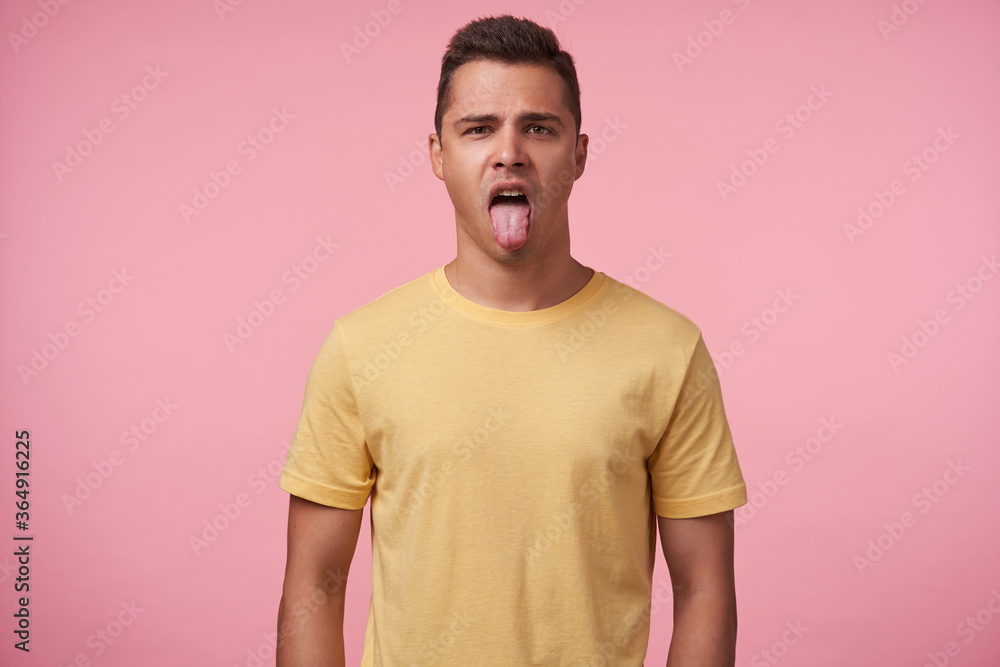 Studio shot of young pretty brown haired male sticking out his tongue while looking at camera, keeping hands along body while posing over pink background Studio shot of young pretty brown haired male sticking out his tongue while looking at camera, keeping hands along body while posing over pink background