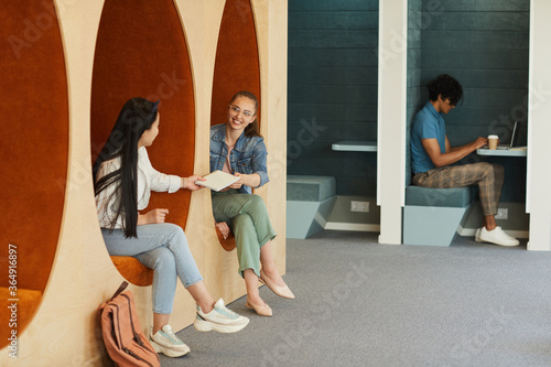 Smiling student girl passing lecture notes to group mate while they sitting in cozy university hall