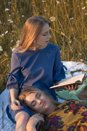 Two female friends recreating in field and have picnic. Sitting and reading a book. Relax in summer outdoor