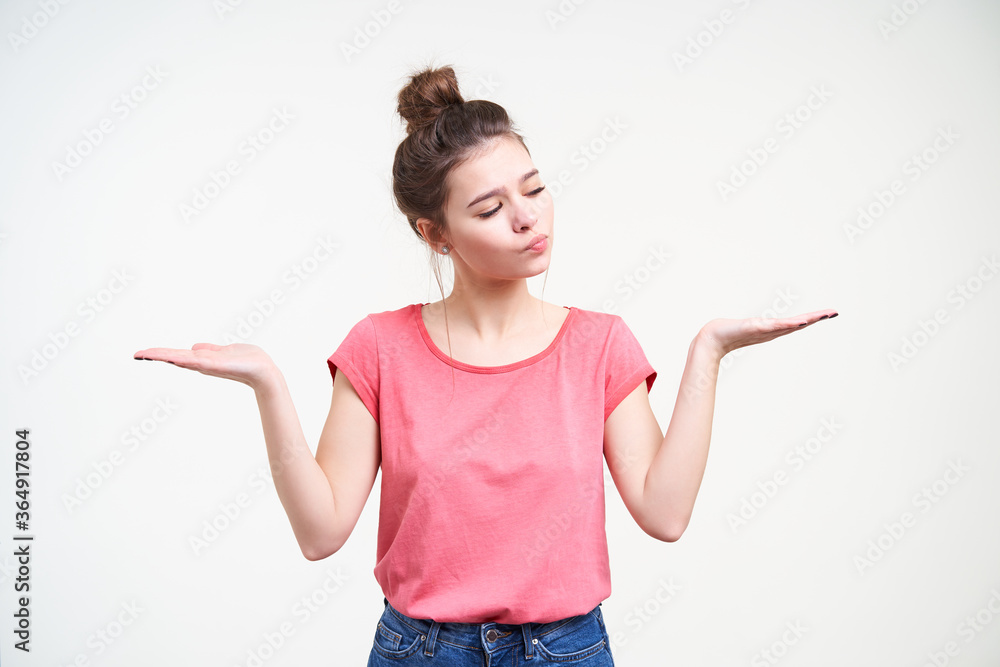 Puzzled young pretty woman wearing her brown hair in knot while posing over white background, keeping palms raised and looking pensively on one of it Puzzled young pretty woman wearing her brown hair in knot while posing over white background, keeping palms raised and looking pensively on one of it