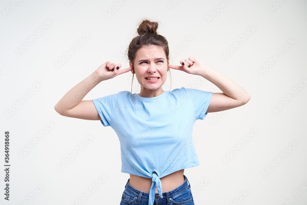 Displeased young pretty brunette female with bun hairstyle frowning her face and covering ears while avoiding loud sounds, standing over white background Displeased young pretty brunette female with bun hairstyle frowning her face and covering ears while avoiding loud sounds, standing over white background