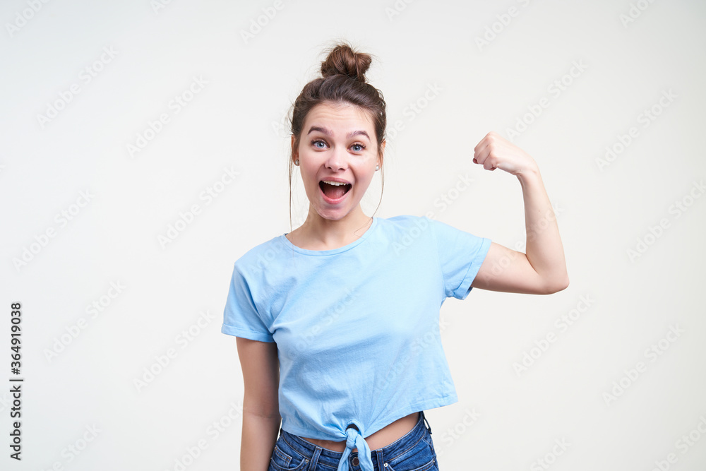 Excited young attractive dark haired woman with bun hairstyle keeping her hand raised while demonstrating her strong biceps, isolated over white background Excited young attractive dark haired woman with bun hairstyle keeping her hand raised while demonstrating her strong biceps, isolated over white background