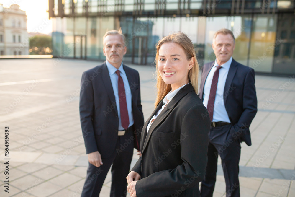 Happy beautiful business woman wearing office suit, standing outdoors and looking at camera. Male business colleagues standing behind. Business team concept Happy beautiful business woman wearing office suit, standing outdoors and looking at camera. Male business colleagues standing behind. Business team concept